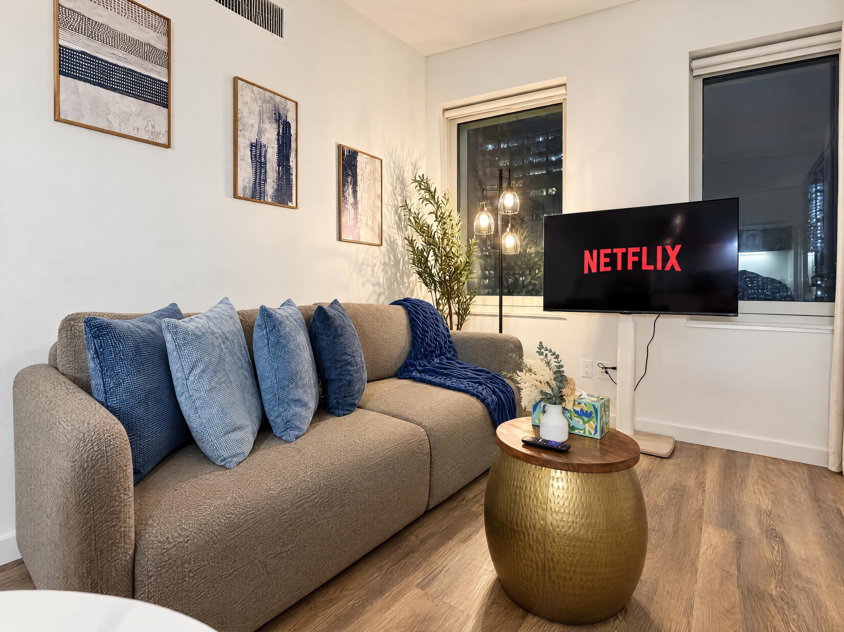 A living room with a beige sofa, blue cushions, a round gold coffee table, and a flat-screen TV displaying 'NETFLIX'.