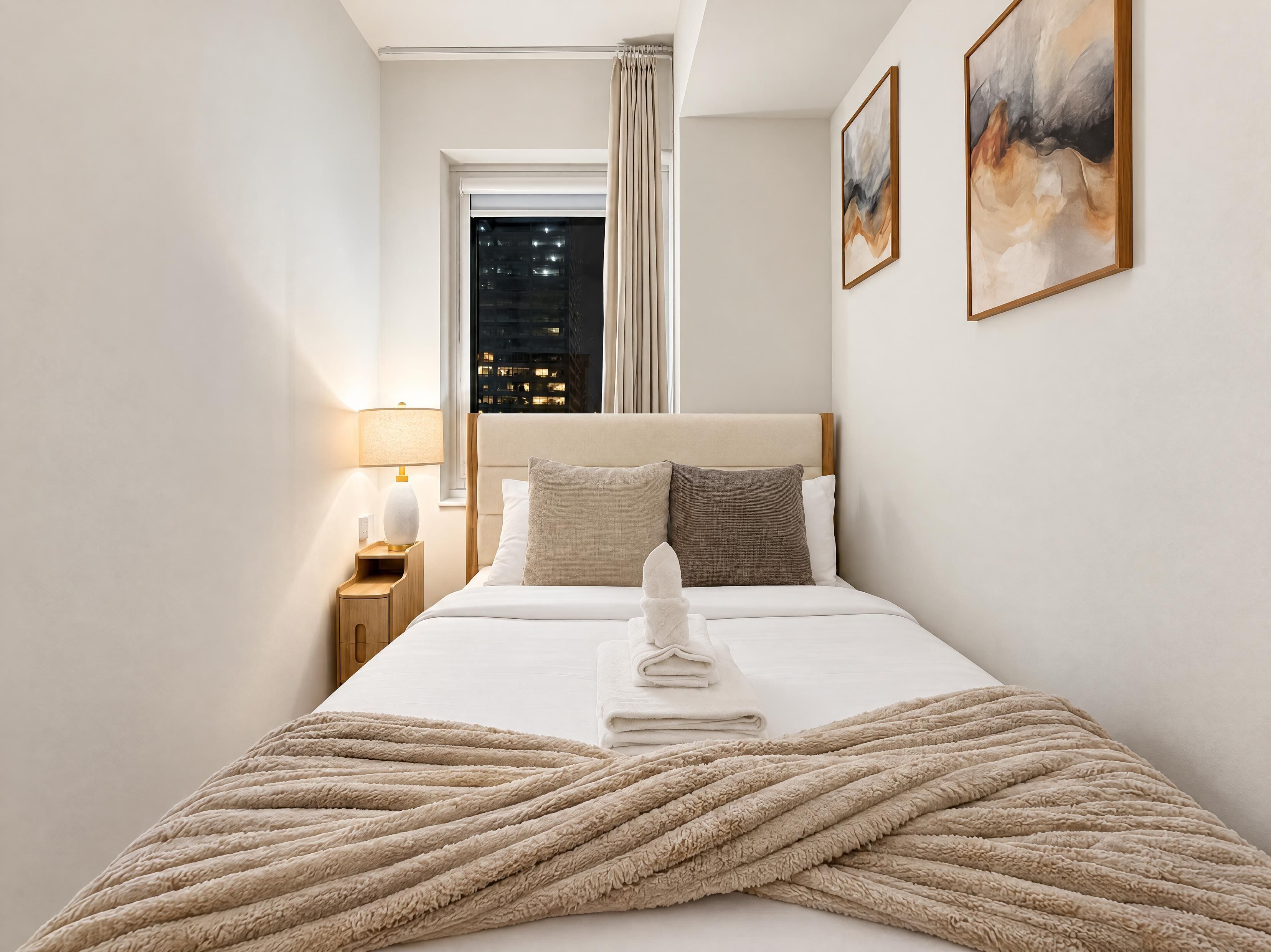 Une chambre d’hôtel avec un lit, une table de chevet, une lampe, une fenêtre avec des rideaux et un tableau au mur.