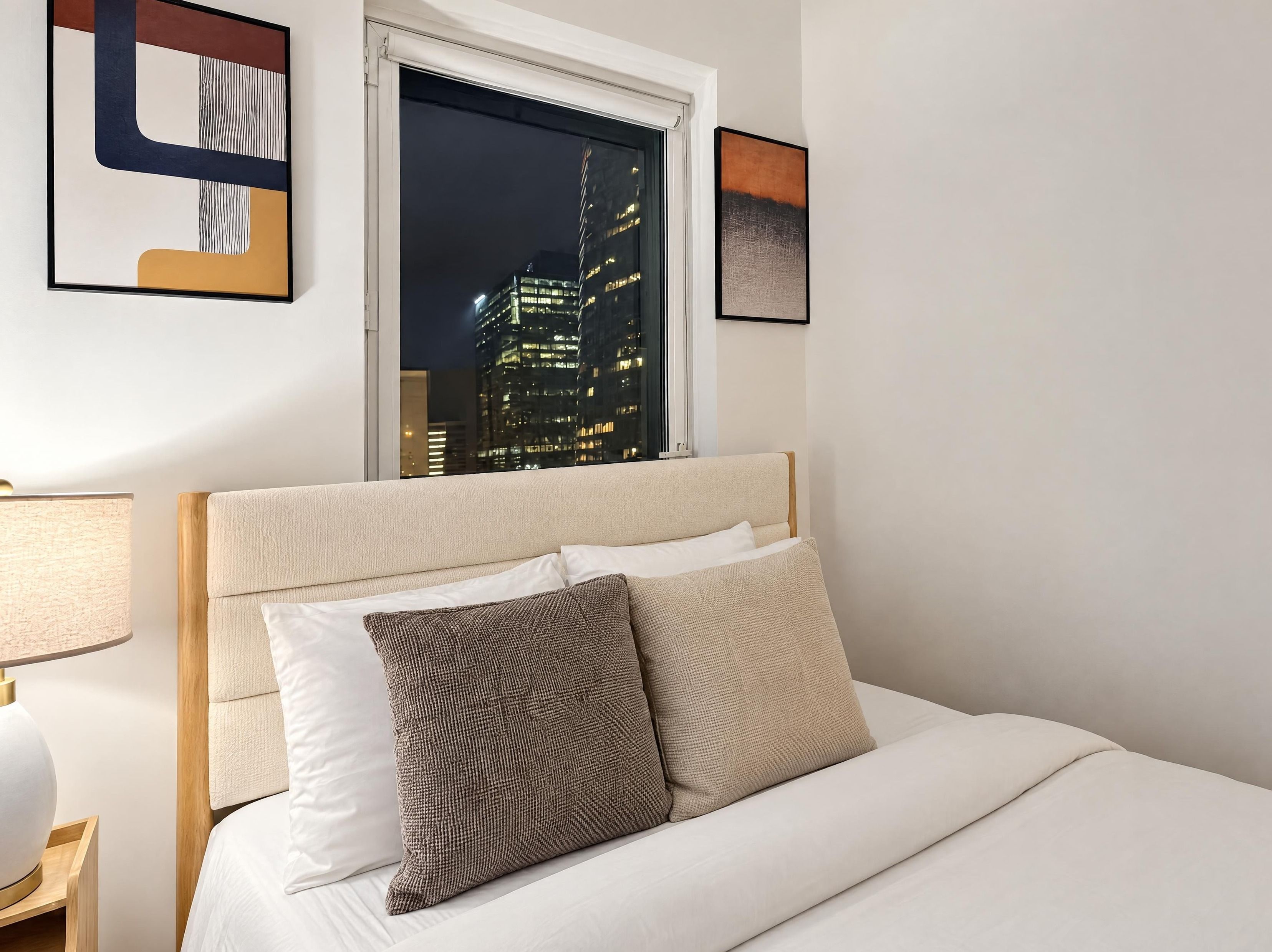 Una camera d'albergo con un letto, vista notturna sulla città e un quadro astratto incorniciato al muro.