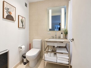A modern bathroom with a toilet, sink, and a mirror. There are towels and a trash bin.