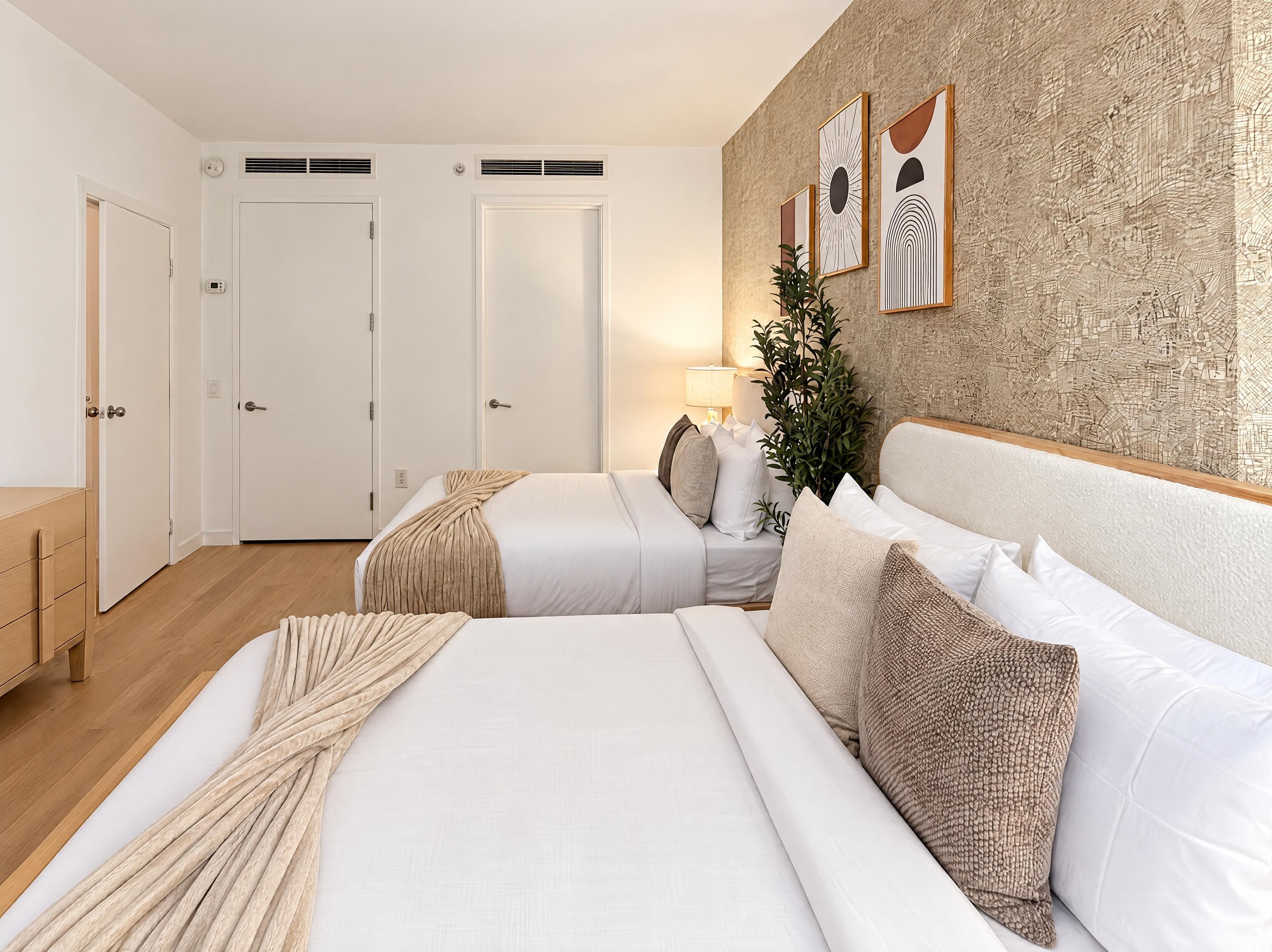 A hotel room with two beds, a wooden floor, a wall with abstract art, and a plant.