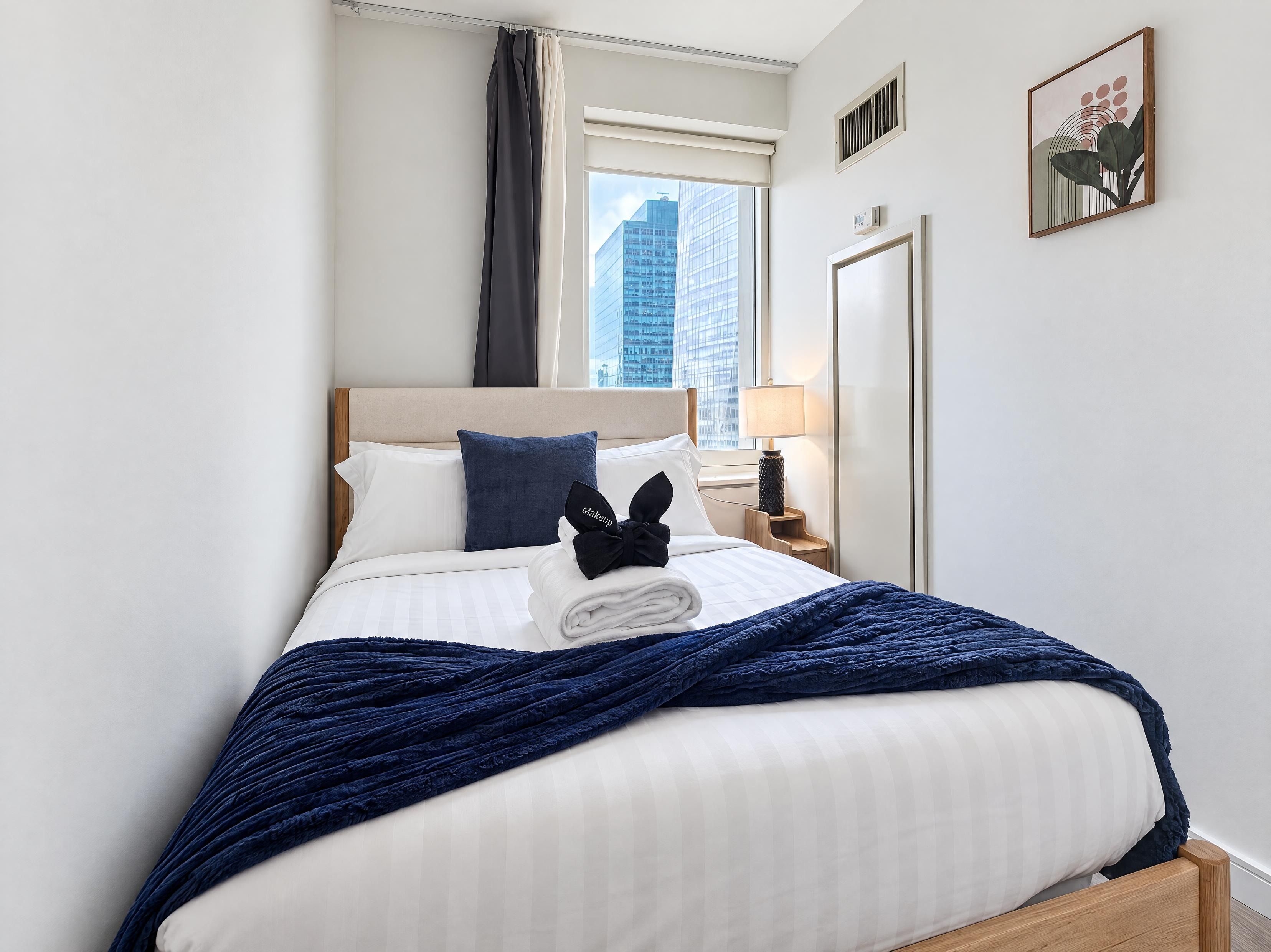 A hotel room with a bed, a nightstand, a lamp, a window with a view of buildings, and a wall-mounted artwork.