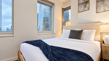 Una camera d'hotel con un letto grande, due finestre, vista sulla città e un comodino con una lampada.