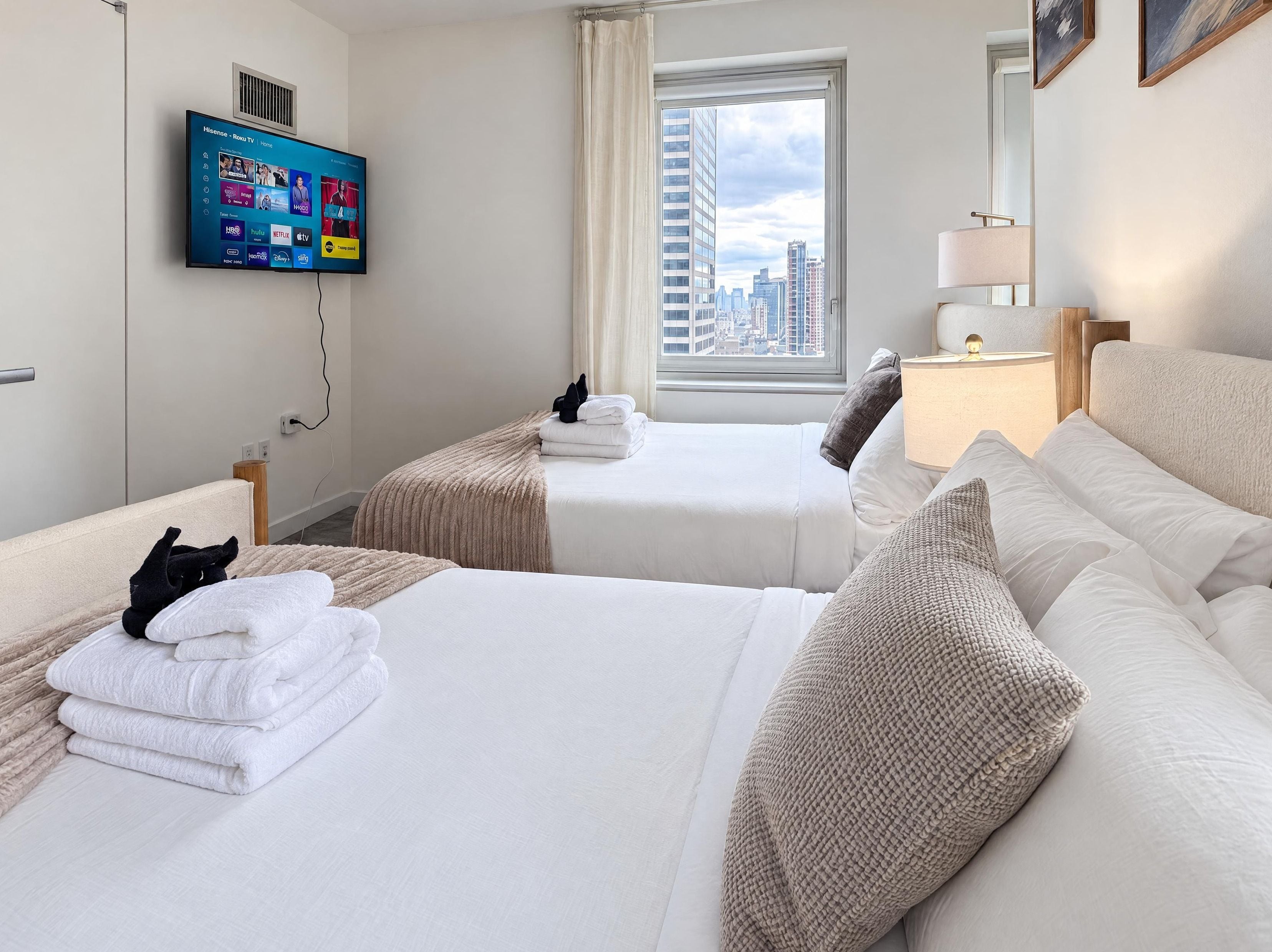 Habitación de hotel con dos camas, un televisor instalado en la pared, una ventana con vista a la ciudad y una mesita de noche con lámpara.