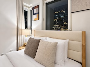 A hotel room with a bed, a nightstand, a lamp, and a window with a cityscape view.