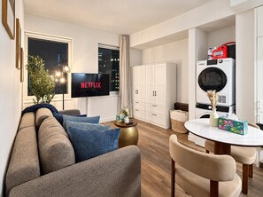 A modern living room with a sofa, a television displaying 'NETFLIX,' a dining area with a table and chairs, and a kitchen area with a washing machine and cabinets.
