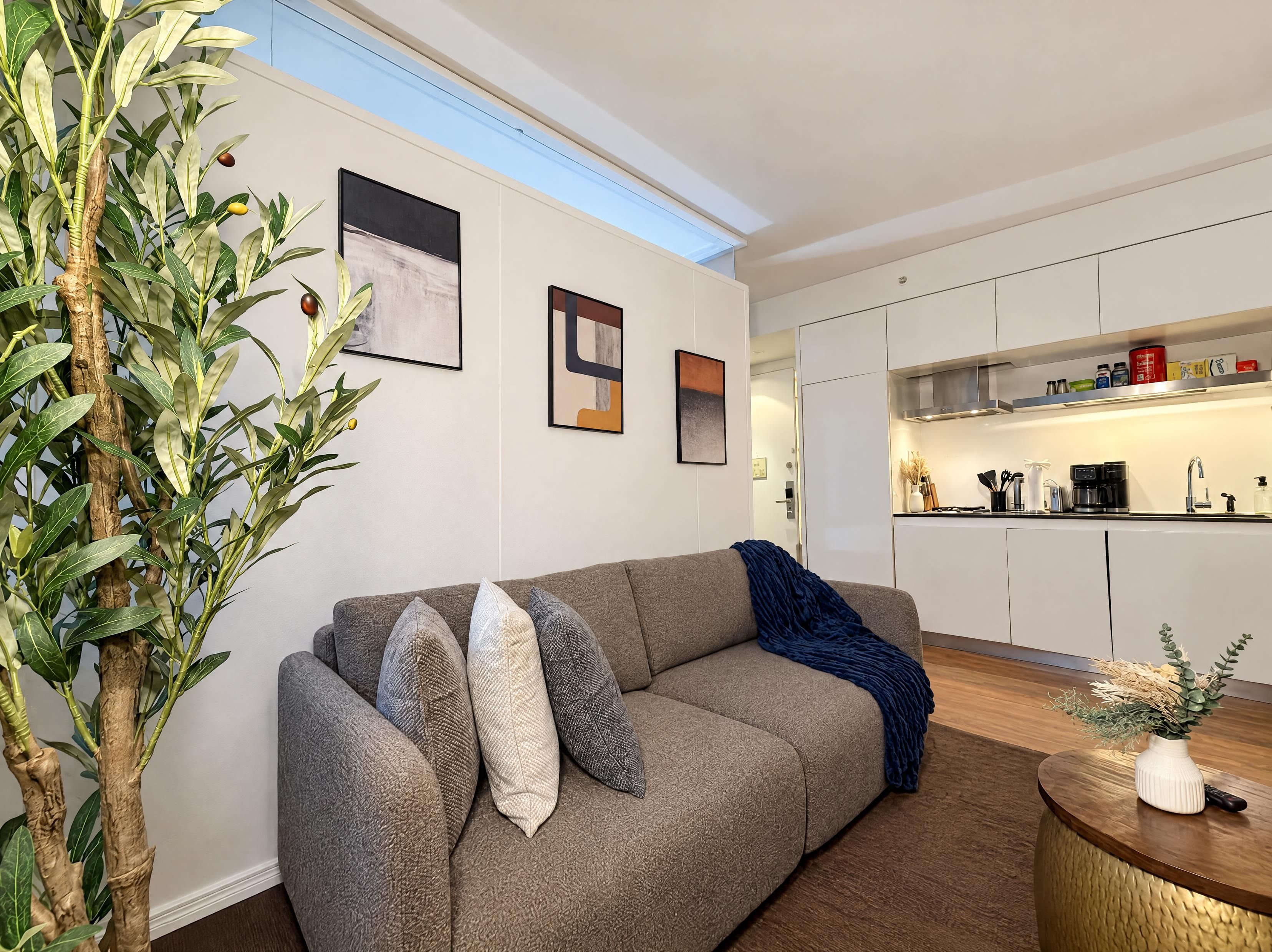 A modern living room with a grey sofa, decorative pillows, a coffee table with a vase, and a kitchen in the background.
