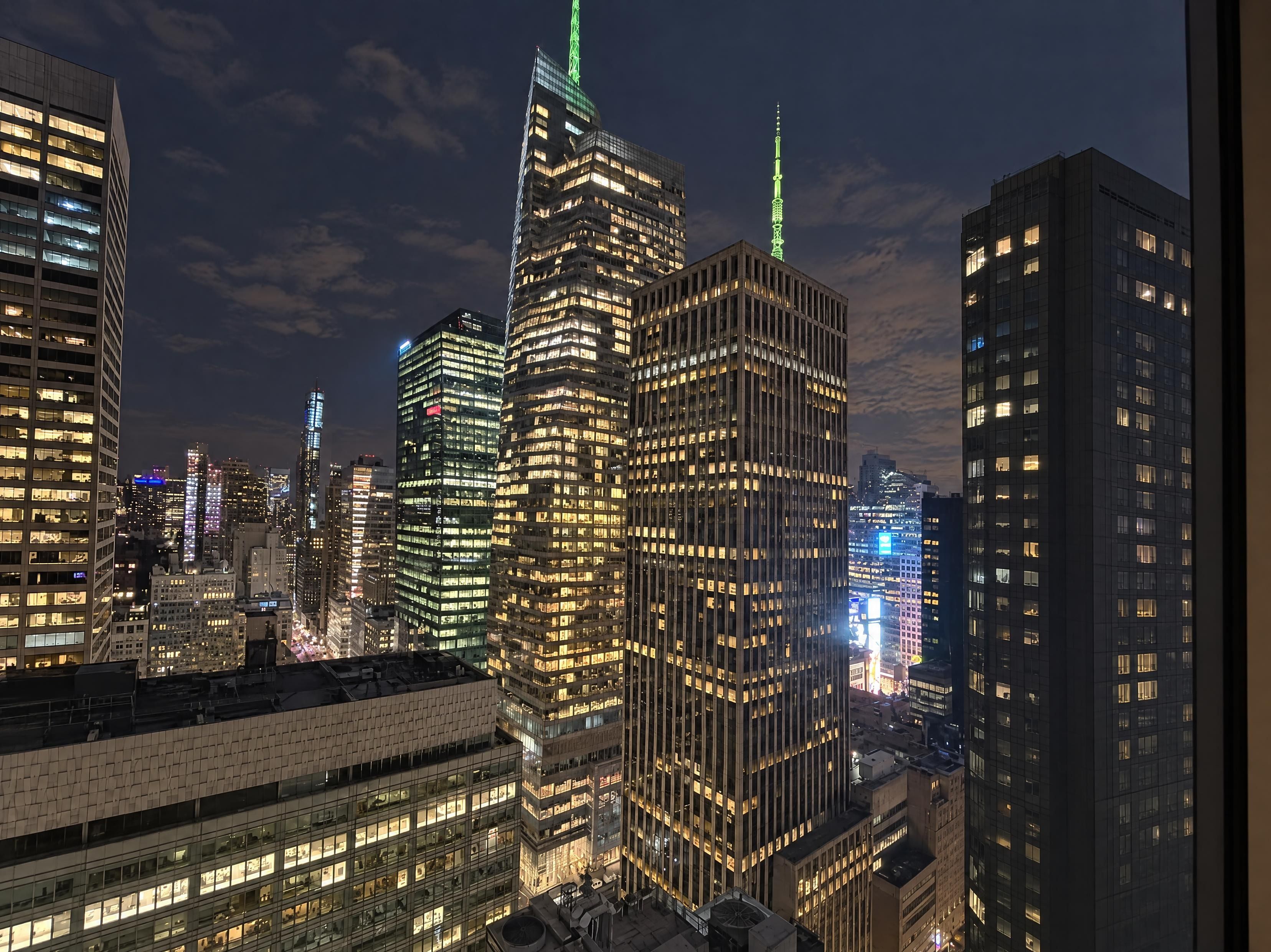 Paesaggio urbano notturno con grattacieli, tra cui un imponente grattacielo con guglia illuminata di verde.
