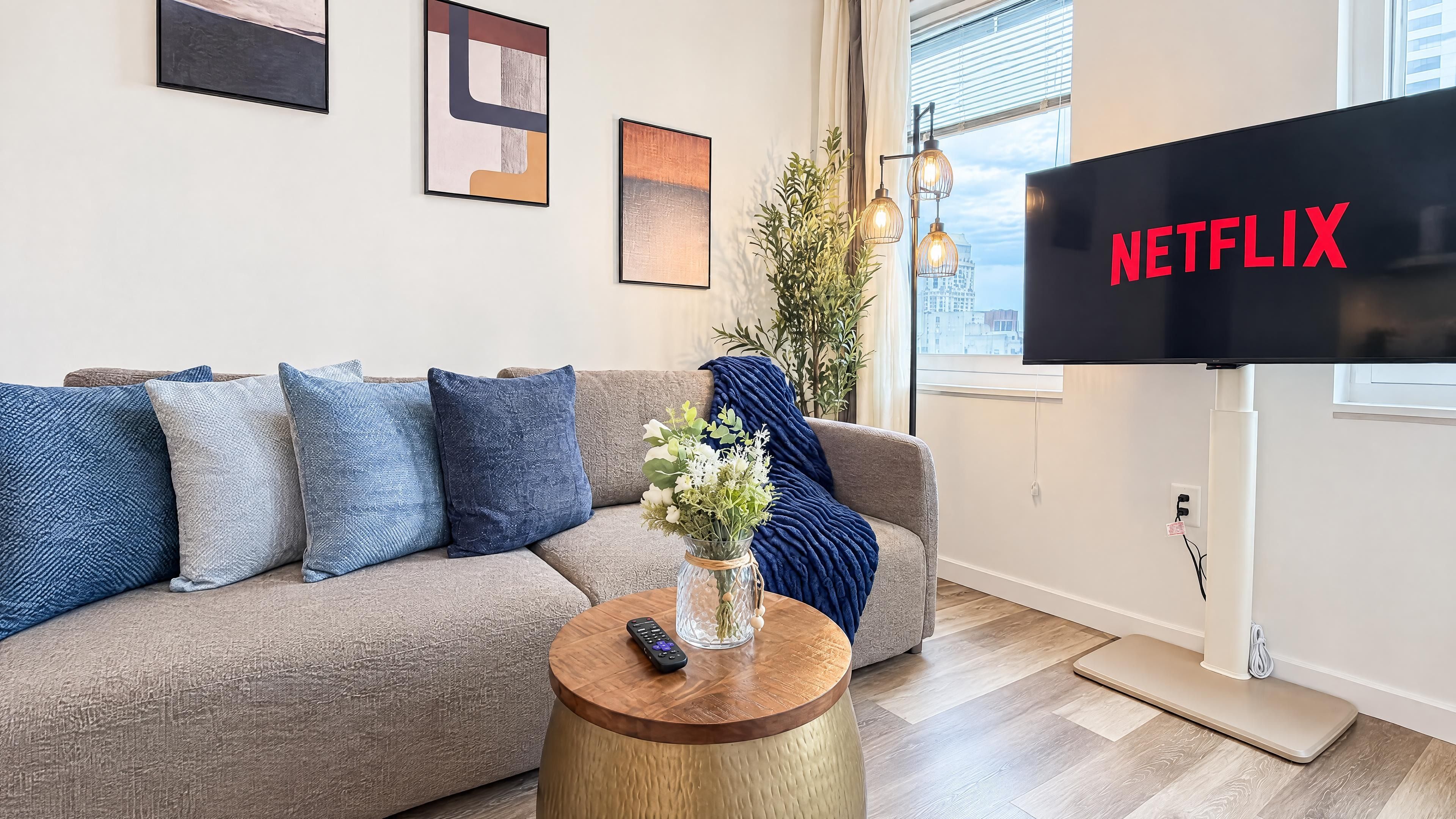 A living room with a grey sofa, blue cushions, a vase with flowers, a wooden coffee table, and a TV displaying 'NETFLIX'.