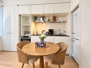 A compact kitchen with white cabinets, a round wooden table, wicker chairs, and a coffee maker.