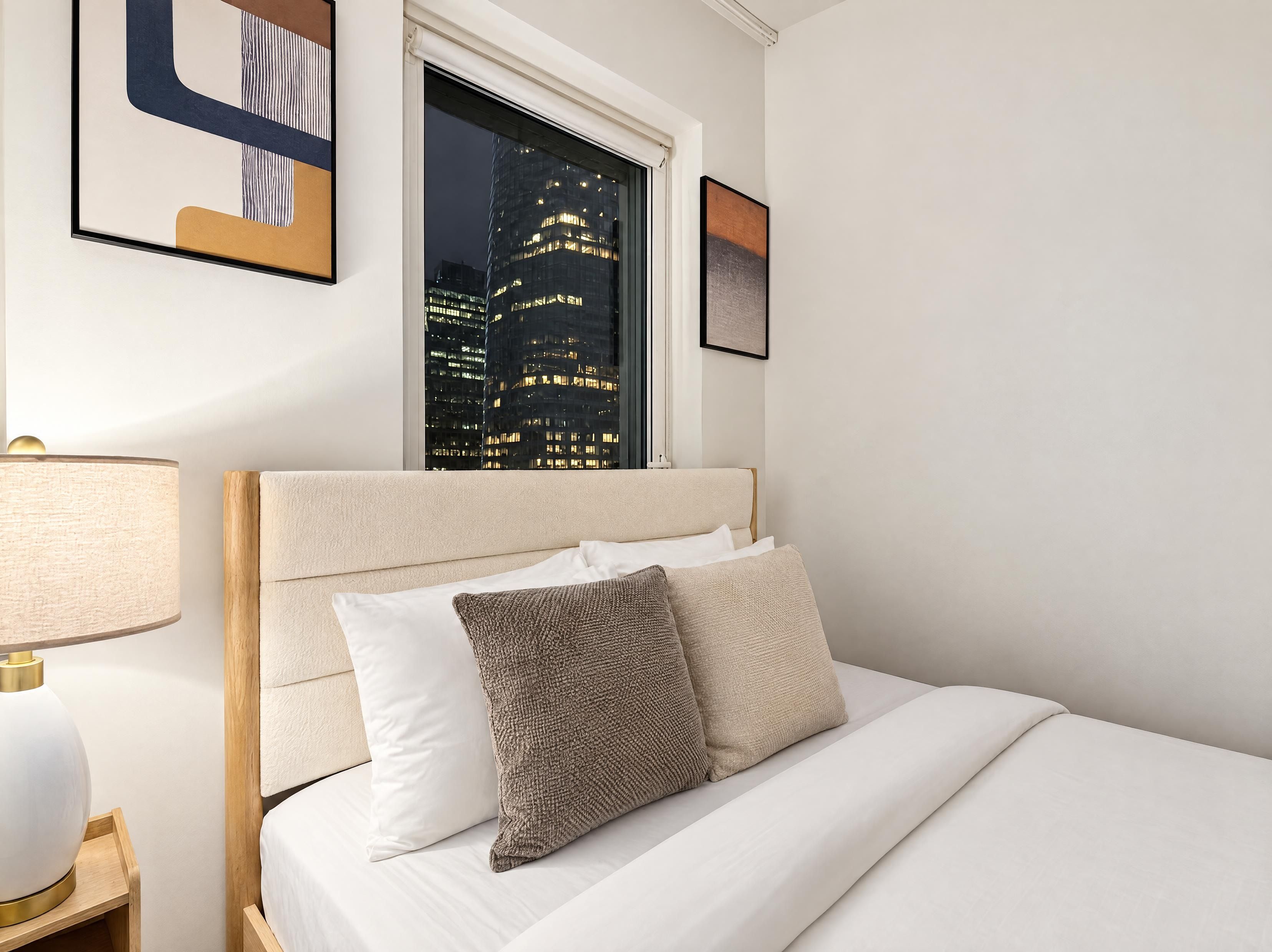 A hotel room with a bed, a nightstand, a lamp, a window with a city view, and framed artwork on the wall.