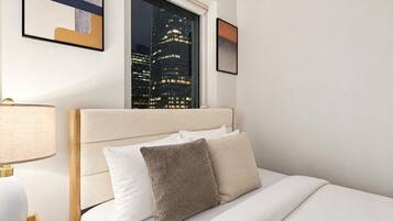 A hotel room with a bed, a nightstand, a lamp, a window with a city view, and framed artwork on the wall.