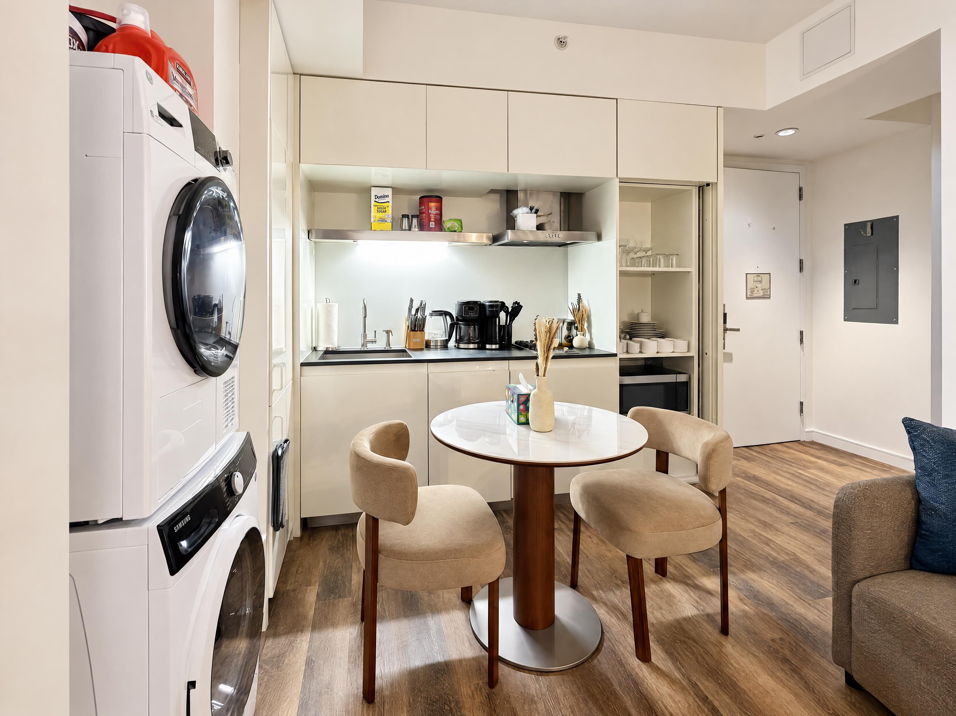 A compact living space with a washing machine, a dining table with chairs, a kitchen area with a coffee maker, and a sofa.