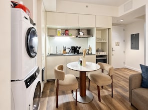 A compact living space with a washing machine, a dining table with chairs, a kitchen area with a coffee maker, and a sofa.