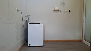 A small, enclosed laundry area with a white washing machine, a hose, and a power outlet on the wall.