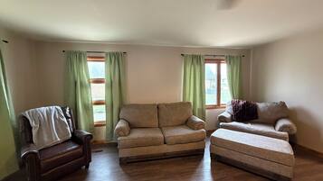 Ein Wohnzimmer mit einem braunen Ledersessel, einem beigen Sofa und einem passenden Hocker. Es gibt zwei Fenster mit grünen Vorhängen.