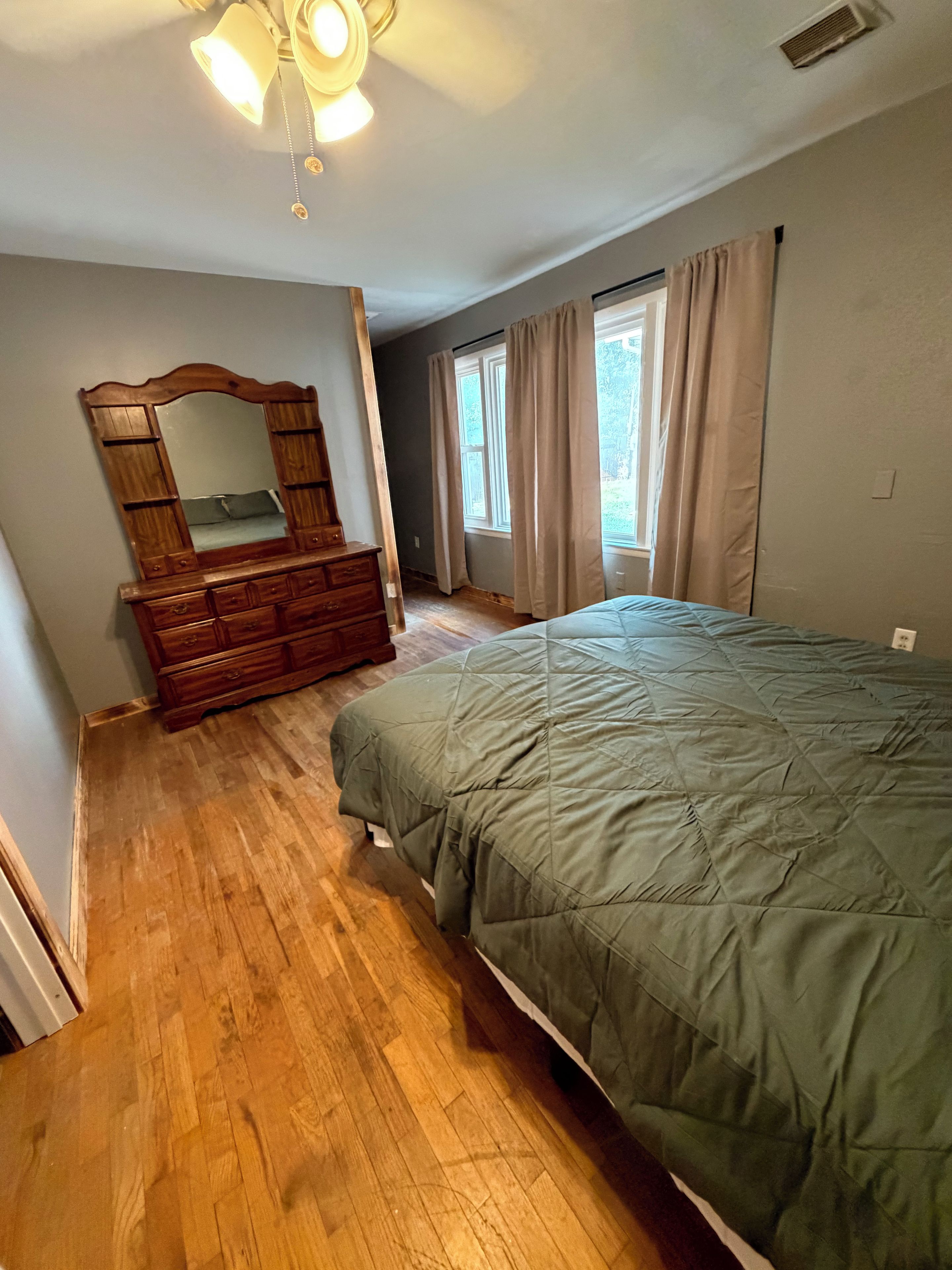 Une chambre à coucher avec une commode en bois, un lit recouvert d’une courtepointe verte, un ventilateur de plafond et une fenêtre avec des rideaux.