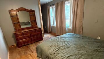 Ein Schlafzimmer mit einem hölzernen Schminktisch, einem Bett mit grüner Tagesdecke, einem Deckenventilator und einem Fenster mit Vorhängen.