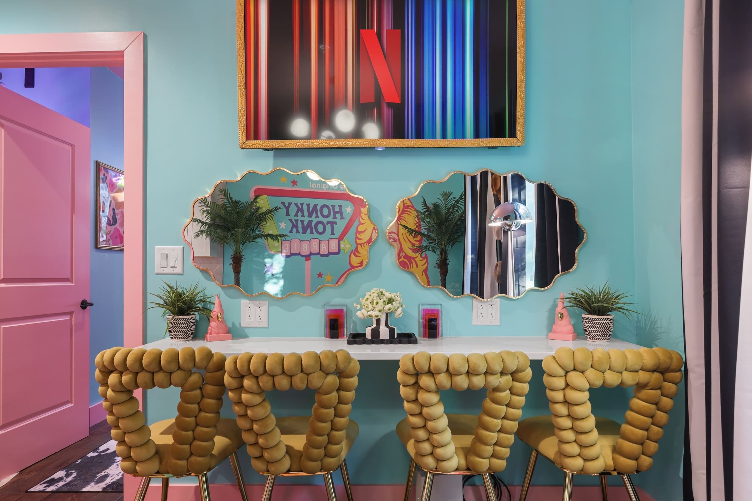 Une pièce aux murs de couleur turquoise, avec une porte rose et une table blanche entourée de quatre chaises jaunes. On y trouve deux miroirs muraux dotés de cadres décoratifs, l’un portant l’inscription « HONEY » et l’autre l’inscription « BARBIE ».