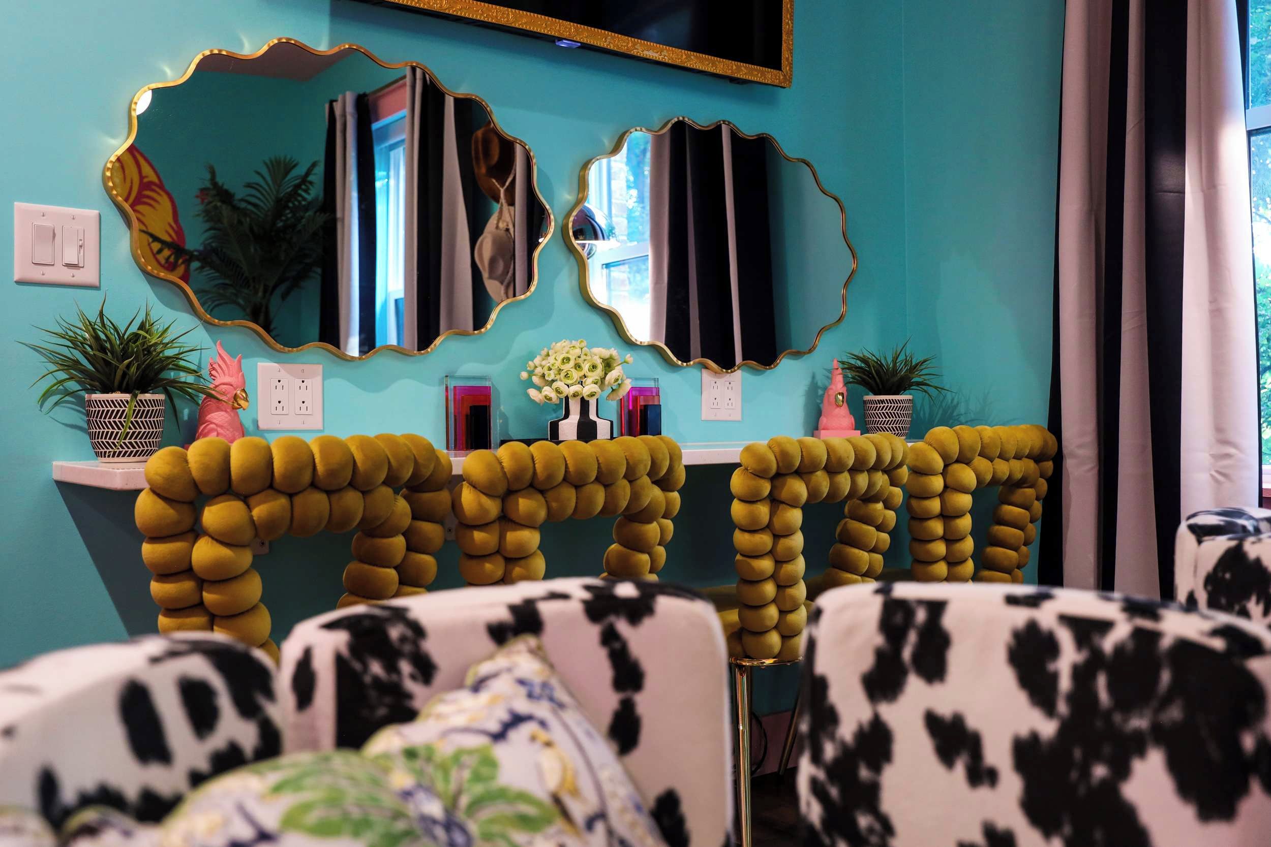 Una habitación con paredes color turquesa, dos espejos grandes con marcos dorados, una consola blanca con un florero y un sofá con estampado blanco y negro.