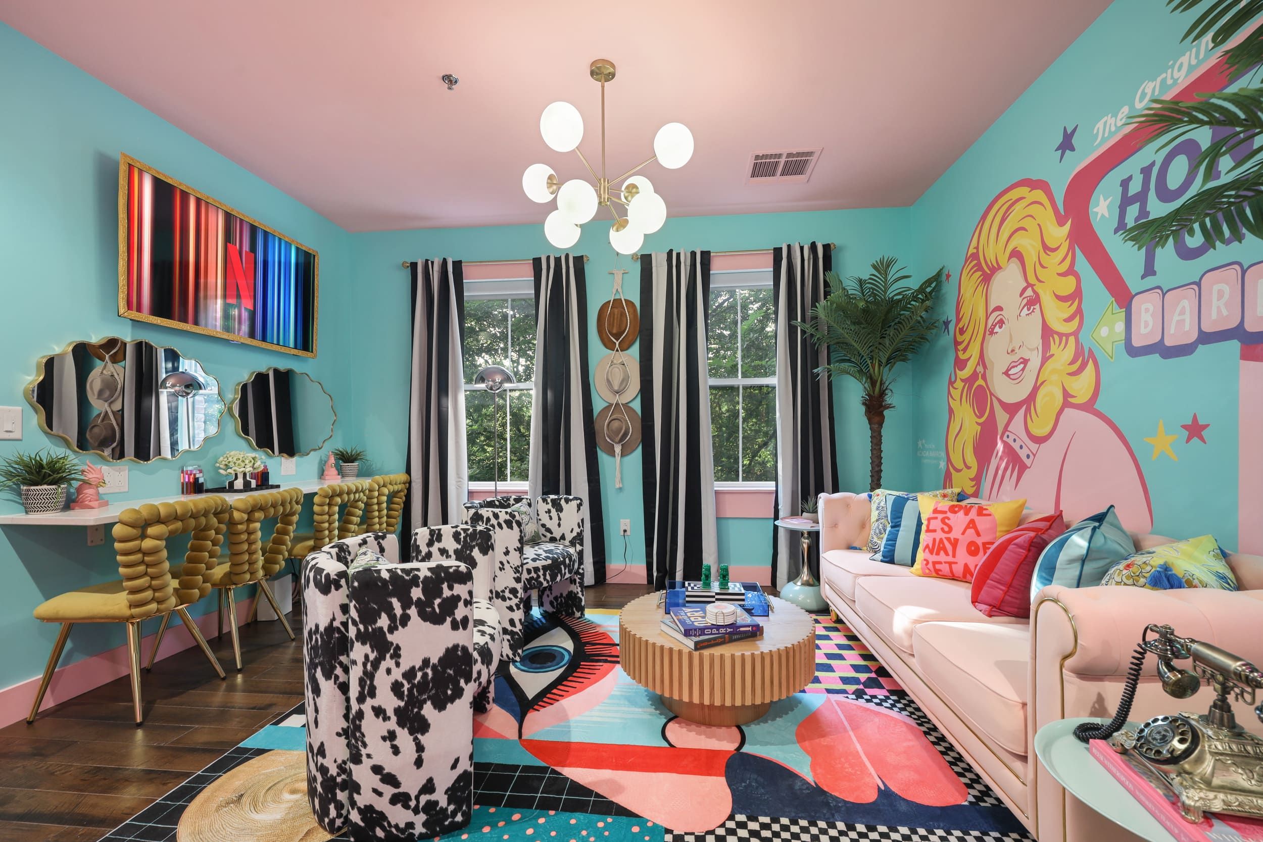 Un salon lumineux et accueillant, avec un mur de couleur turquoise, un grand miroir, un luminaire au plafond et un tapis aux couleurs vives.