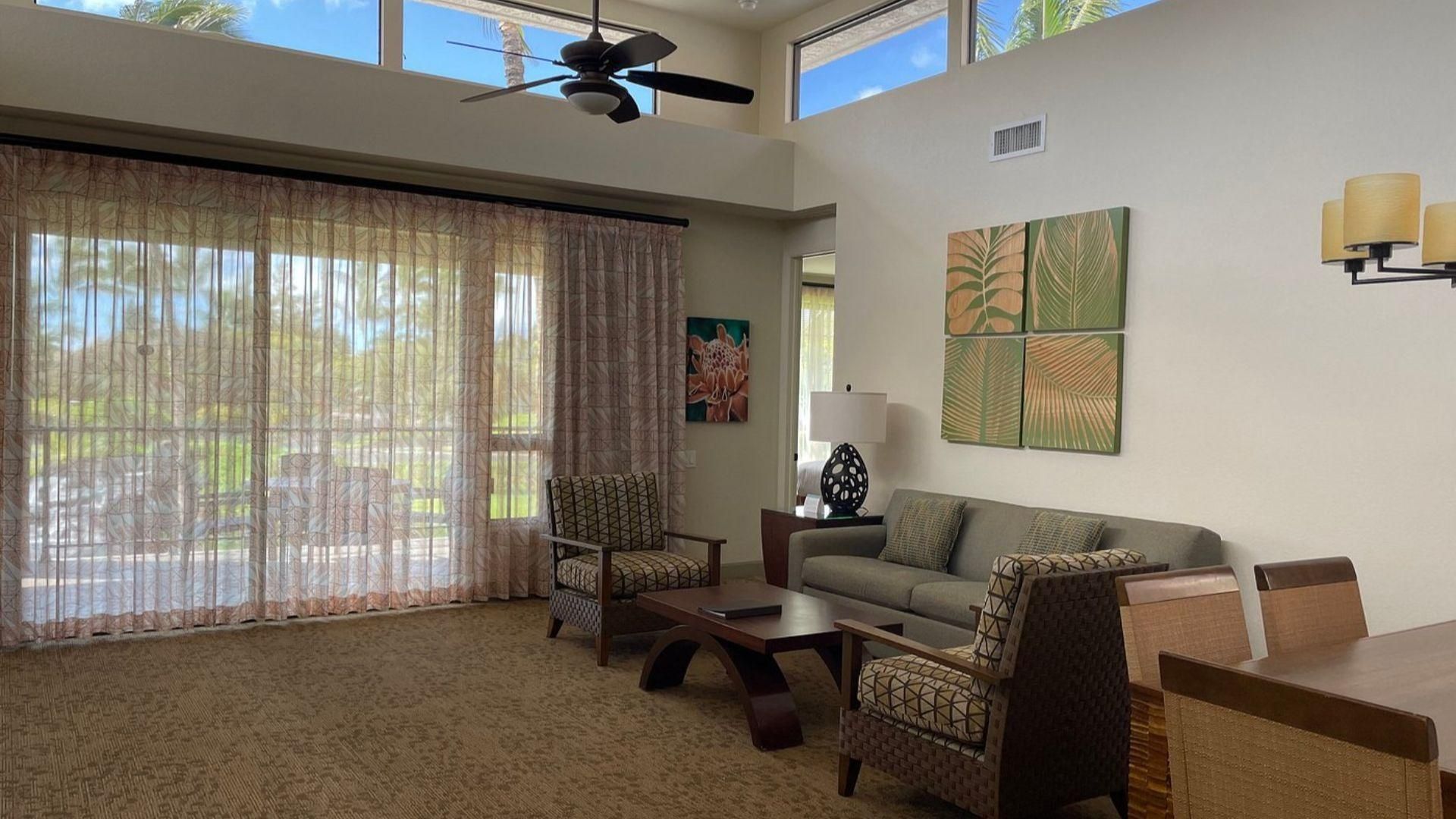 A spacious living room with a large window, a ceiling fan, a sofa, two armchairs, a coffee table, and wall art.