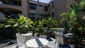 Eine Terrasse im Freien mit weißen Tischen und Stühlen, umgeben von üppiger tropischer Bepflanzung und einem Grill.