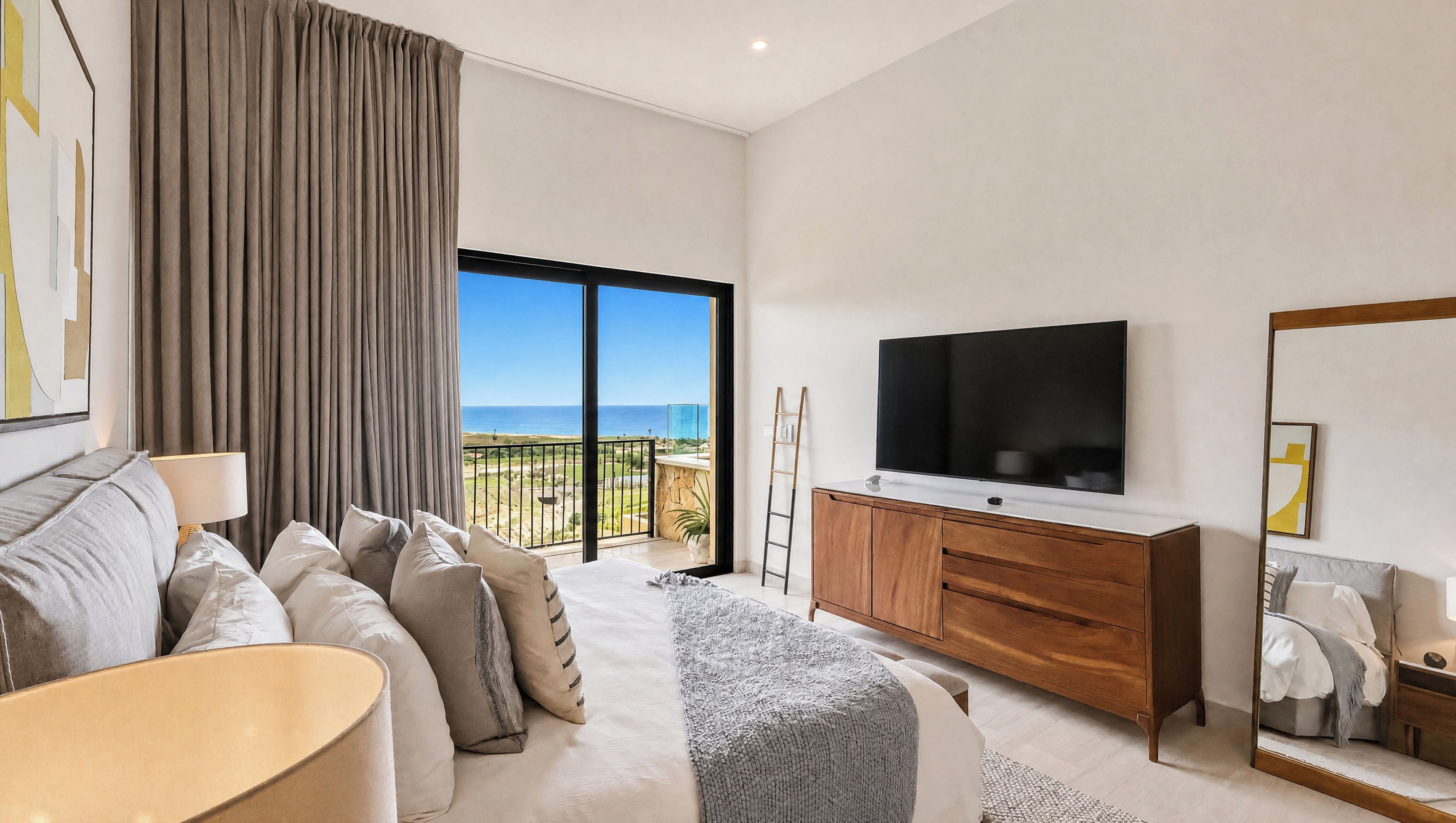 A modern bedroom with a large TV, a balcony with a view of the sea, and a cozy seating area.