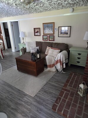 A cozy living room with a brown coffee table, a sofa, and a green nightstand. There are framed pictures on the wall and a door leading to another room. - Adorable 1-bedroom apartment in Conway perfect for your getaway (Conway)