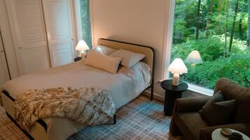 A bedroom with a bed, two bedside lamps, a window with a view of greenery, a sofa, and a small table.