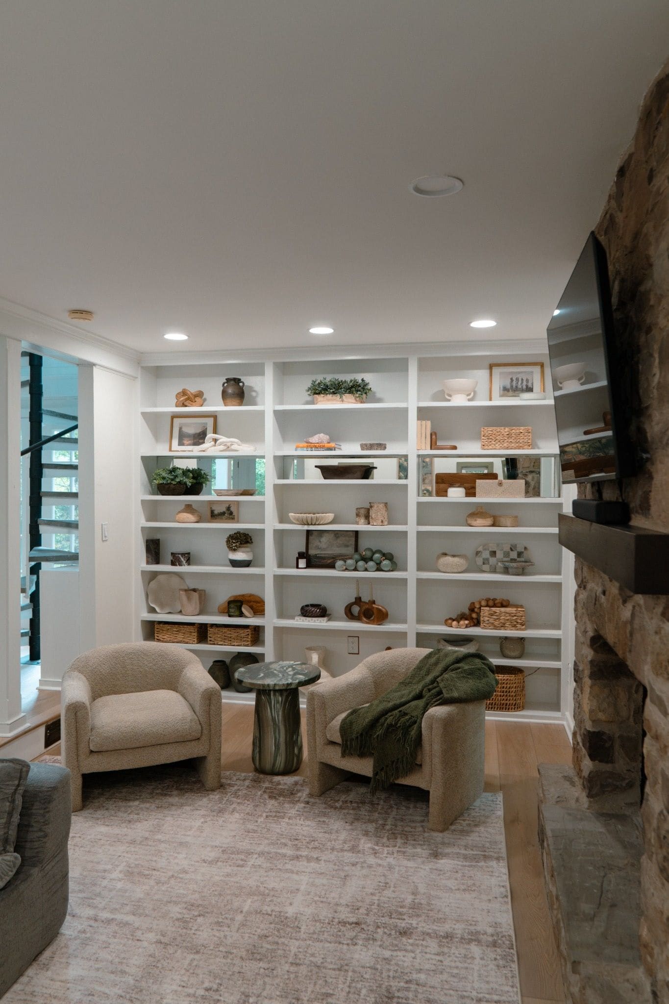 A cozy living room with a fireplace, a bookshelf filled with decorative items, two armchairs, and a coffee table.