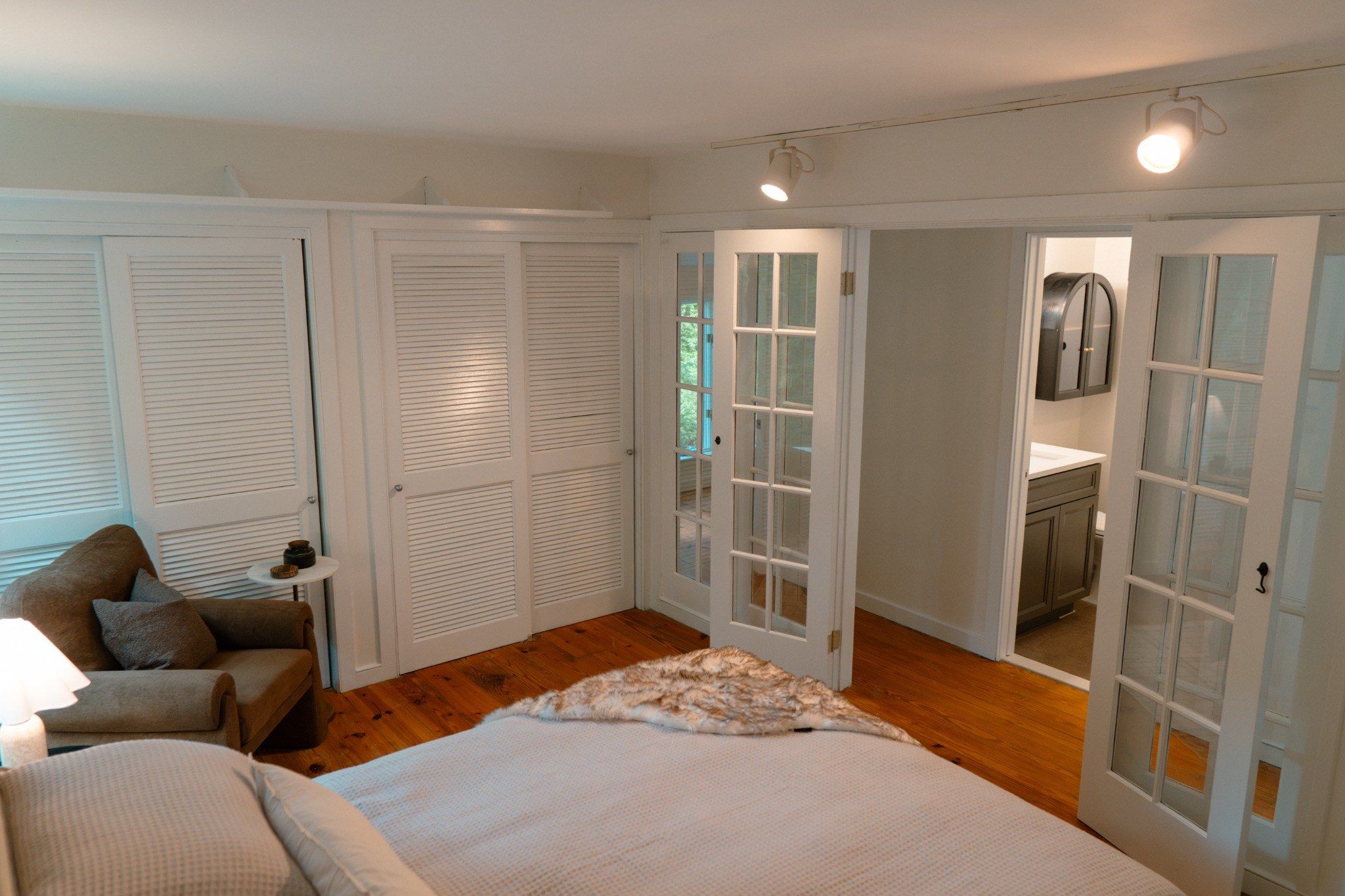 A bedroom with a bed, a nightstand, a lamp, a sofa, and a view of a bathroom through a glass door.