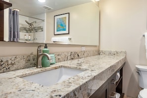 Ein Badezimmer mit einer Arbeitsplatte aus Granit, einem Waschbecken, einer Dusche mit Glastür, einem gerahmten Bild an der Wand und einem Handtuchhalter.