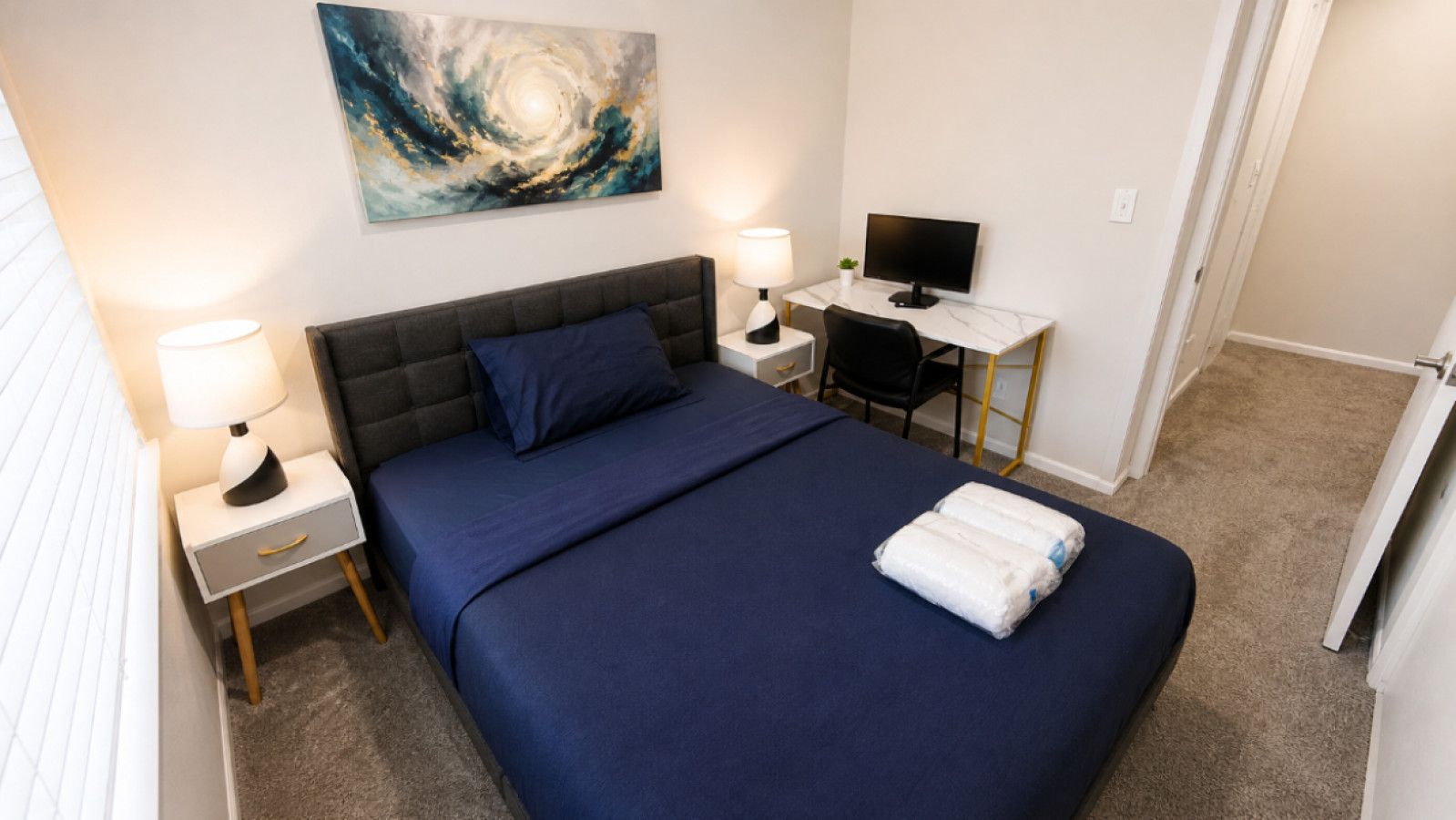 A bedroom with a bed, two bedside tables with lamps, a desk with a computer, and a painting on the wall.