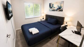 Ein Schlafzimmer mit Bett, Nachttischen, einem Schreibtisch mit Computer, einem Fernseher und einem Fenster mit Jalousien.