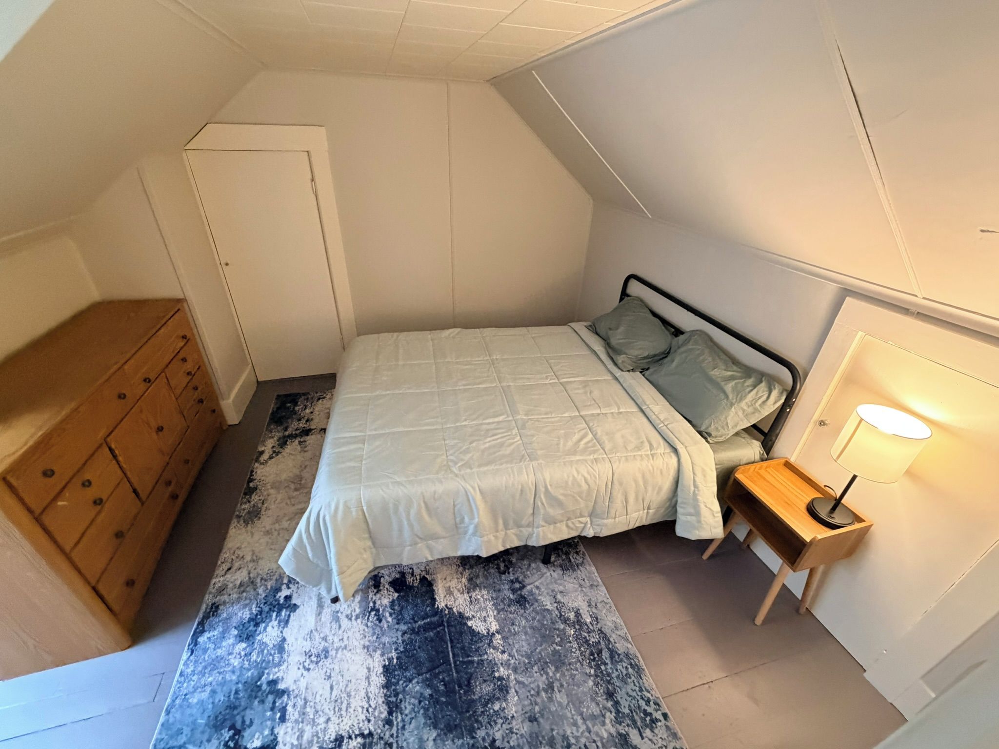 A small, single-bed room with a sloped ceiling, a wooden dresser, a bedside table with a lamp, and a patterned rug.