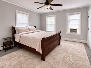 Quarto com cama, mesa de cabeceira, ventilador de teto e três janelas com persianas.