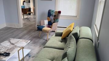 Un salon avec un canapé vert, un fauteuil à motifs et une grande fenêtre avec des stores.