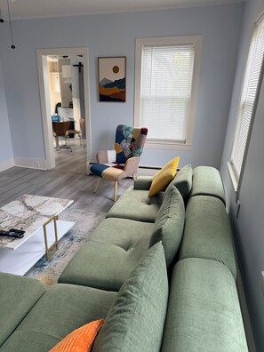 Una sala de estar con un sofá verde, un sillón estampado y un ventanal con persianas.