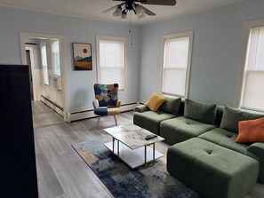 A living room with a green sofa, a patterned armchair, a coffee table, and a ceiling fan.