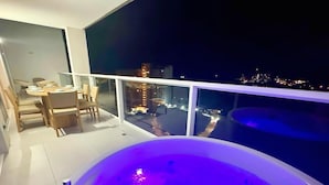 Ein Balkon mit Whirlpool, ein Essbereich mit Stühlen und Tisch sowie ein Blick auf die nächtliche Stadt.