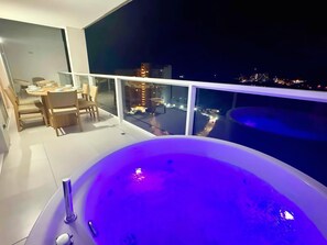 Een balkon met een jacuzzi, een eethoek met stoelen en een tafel, en uitzicht op de stad 's nachts.