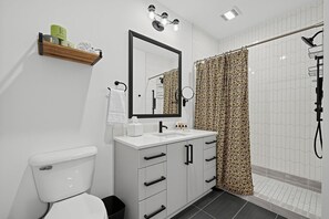 Casa de banho moderna com sanita, toucador com espelho, chuveiro com cortina estampada e um porta-toalhas.