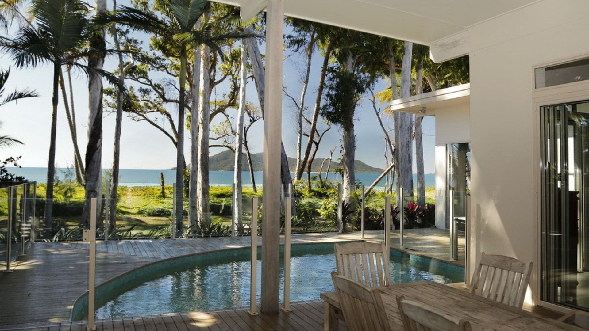 A pool area with a view of the ocean and mountains, surrounded by palm trees and a wooden deck.