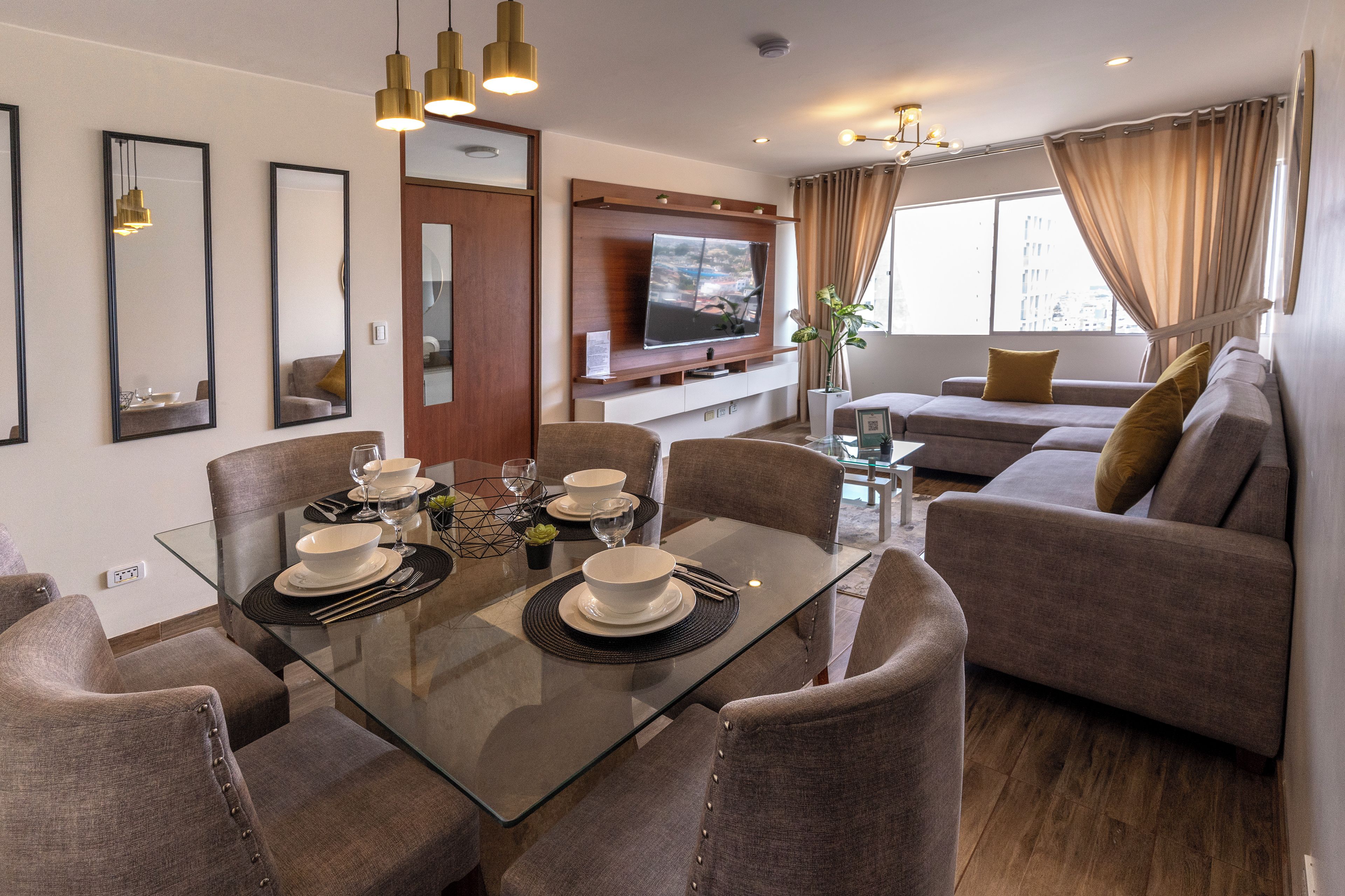 Un salon moderne comprenant un coin repas, un ensemble de canapés, un téléviseur à écran plat fixé au mur et une grande fenêtre avec des rideaux.