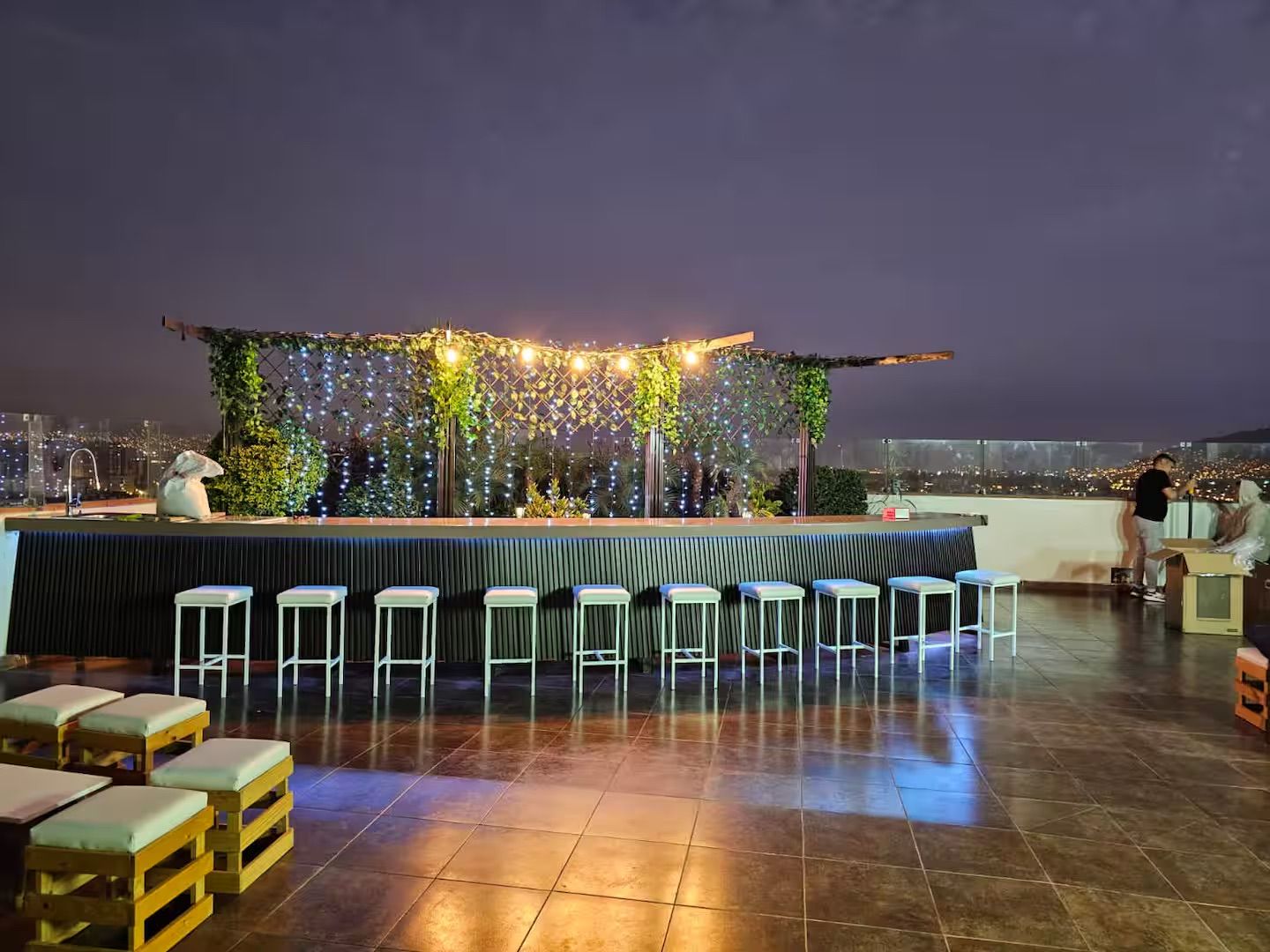 A rooftop bar with a long counter, multiple stools, and a view of the cityscape.