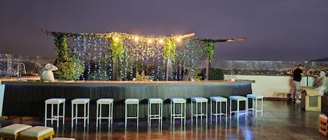 A rooftop bar with a long counter, multiple stools, and a view of the cityscape.