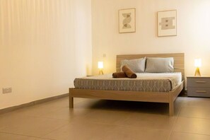 A neatly made bed with a patterned bedspread, two pillows, and a rolled towel on the side. There are two bedside tables with lamps, and the room has a clean, minimalist design. - Spacious 3BR home with Balcony, WIFI & AC (Għarb)
