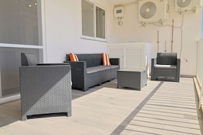 A room with a grey wicker sofa, a matching armchair, and a small table. There is a window and an air conditioning unit on the wall. - Spacious 3BR home with Balcony, WIFI & AC (Għarb)