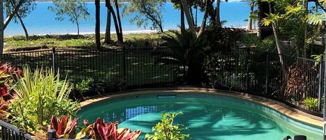 A swimming pool with a view of the sea, surrounded by tropical plants and a metal fence.